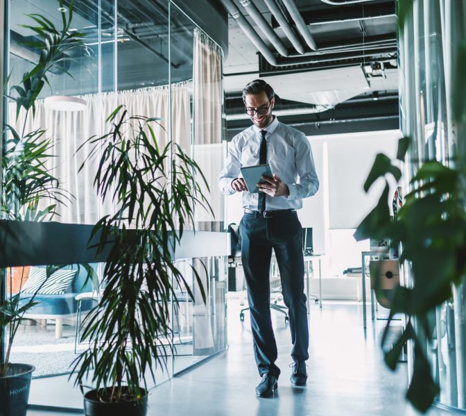 Businessman walking through company and using tablet. Full length.
